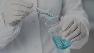 A scientist wearing gloves and a lab coat carefully pours a blue liquid from a test tube into a beaker, representing the Specialty Materials sector.