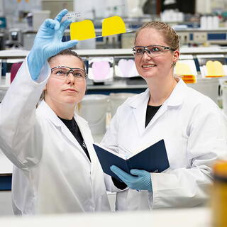Two scientists in lab coats and safety glasses examine a bright yellow sample on a glass slide while one takes notes, representing the Specialty Materials sector.