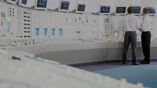 Two men in business attire stand in a spacious control room monitoring systems on screens above complex control panels, representing A&D and Government Services category