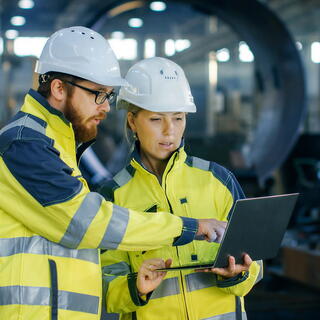 Two workers in hard hats and high-visibility jackets collaborate while reviewing data on a laptop in an industrial facility, representing A&D and Government Services category.