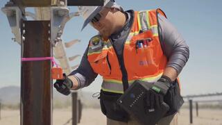 A medium shot of a male construction worker wearing a high-visibility orange safety vest, a white hard hat, sunglasses, and black gloves. He is standing in a desert environment and is inspecting an orange device attached to a metal post with pink flagging tape. In his other hand, he holds a black tablet case that says "SUNSCREEN" on the back. The background shows rows of solar panel support structures stretching into the distance under a clear blue sky.