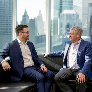 Two American Securities male employees in business casual attire sit on a sofa and converse in a modern office overlooking a city skyline.