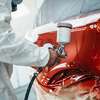A close-up shot of a worker in a white protective hazmat suit and respirator mask using a professional spray gun to apply a glossy, metallic red finish to a vehicle body. The surrounding areas of the car are meticulously covered with masking tape and plastic sheeting to protect them from overspray. The lighting highlights the smooth, reflective quality of the fresh paint in a clean, industrial workshop setting.
