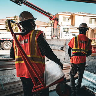Two FBM construction workers in hard hats and high-visibility safety vests stand among building materials at a residential construction site.