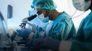 A male scientist in a sterile laboratory setting focuses intently while looking through a high-powered binocular microscope. He is wearing full personal protective equipment, including a teal surgical gown, a matching hairnet, blue gloves, and a face mask. In the foreground and background, other lab personnel in similar attire are partially visible, suggesting a busy, collaborative research or diagnostic environment. The lighting is bright and clinical, emphasizing the precision of the work.
