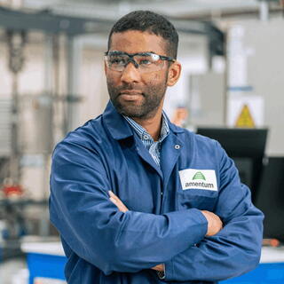 A medium shot of a male professional standing with his arms crossed in an industrial or laboratory environment. He has short dark hair, a light beard, and is wearing clear safety glasses, a patterned button-down shirt, and a blue work jacket featuring an "amentum" logo patch on the left breast. He is looking off to the side with a serious, focused expression against a softly blurred background of technical machinery and equipment.
