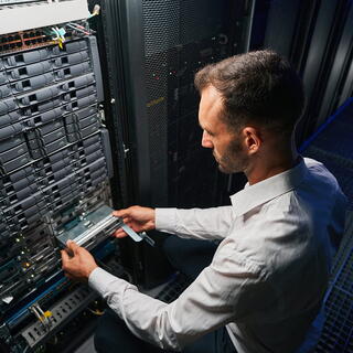A male IT technician in a white shirt installs a hardware component into a dense server rack, representing IT Services sector.