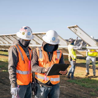 Two SOLV energy workers in safety gear review information on a tablet at a solar panel installation site, representing Power and Energy Sector.