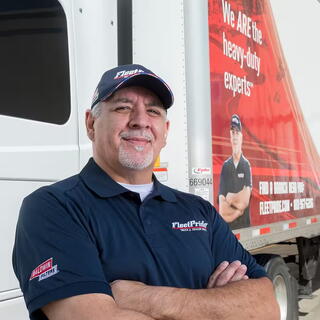 A smiling FleetPride employee stands with his arms crossed in front of a branded company delivery truck, representing the Industrial Products & Services sector.