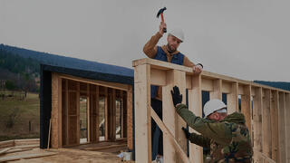 Two construction workers in hard hats collaborate on framing a wooden structure, with one raising a hammer to strike a nail, representing building products sector.